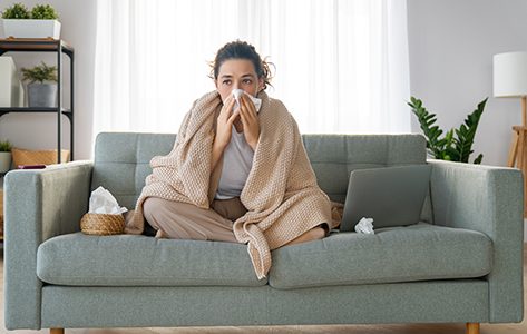 A woman with the flu sits on her sofa, she is wrapped in a blanket and holds tissues.