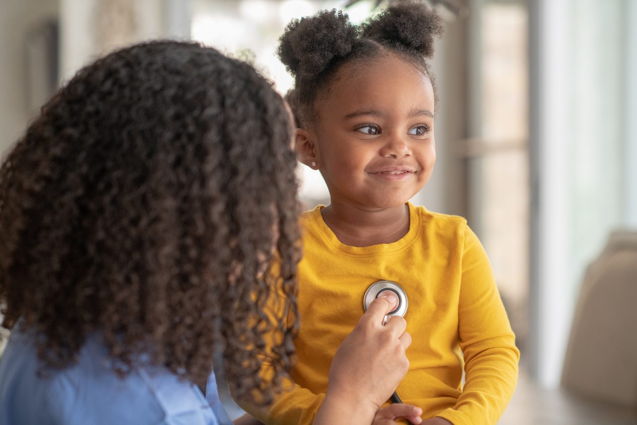 A little girl of African descent is at a medical appointment. She is sitting on an exam table. The female doctor is using a stethoscope to check the little girl's heart rate.