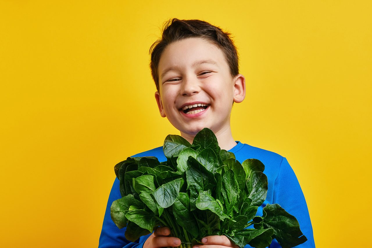 Child happily holding a bunch of spinach on yellow background