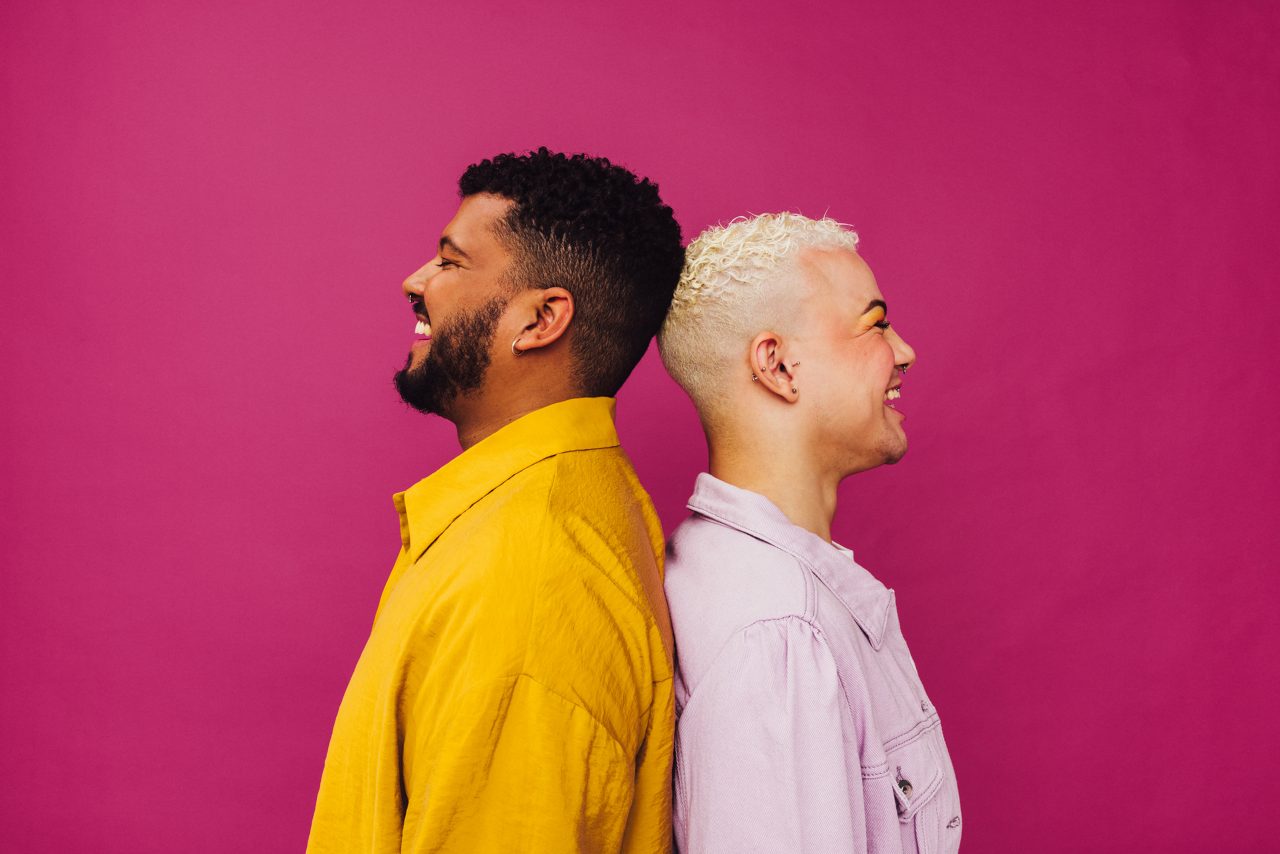 Cheerful gay couple standing together in a studio. Sideview of a beautiful gay couple with piercings standing back to back against a purple background. Young LGBTQ+ couple smiling happily.