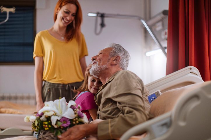Young girl giving her grandpa flowers while visiting hi in hospital with her mother