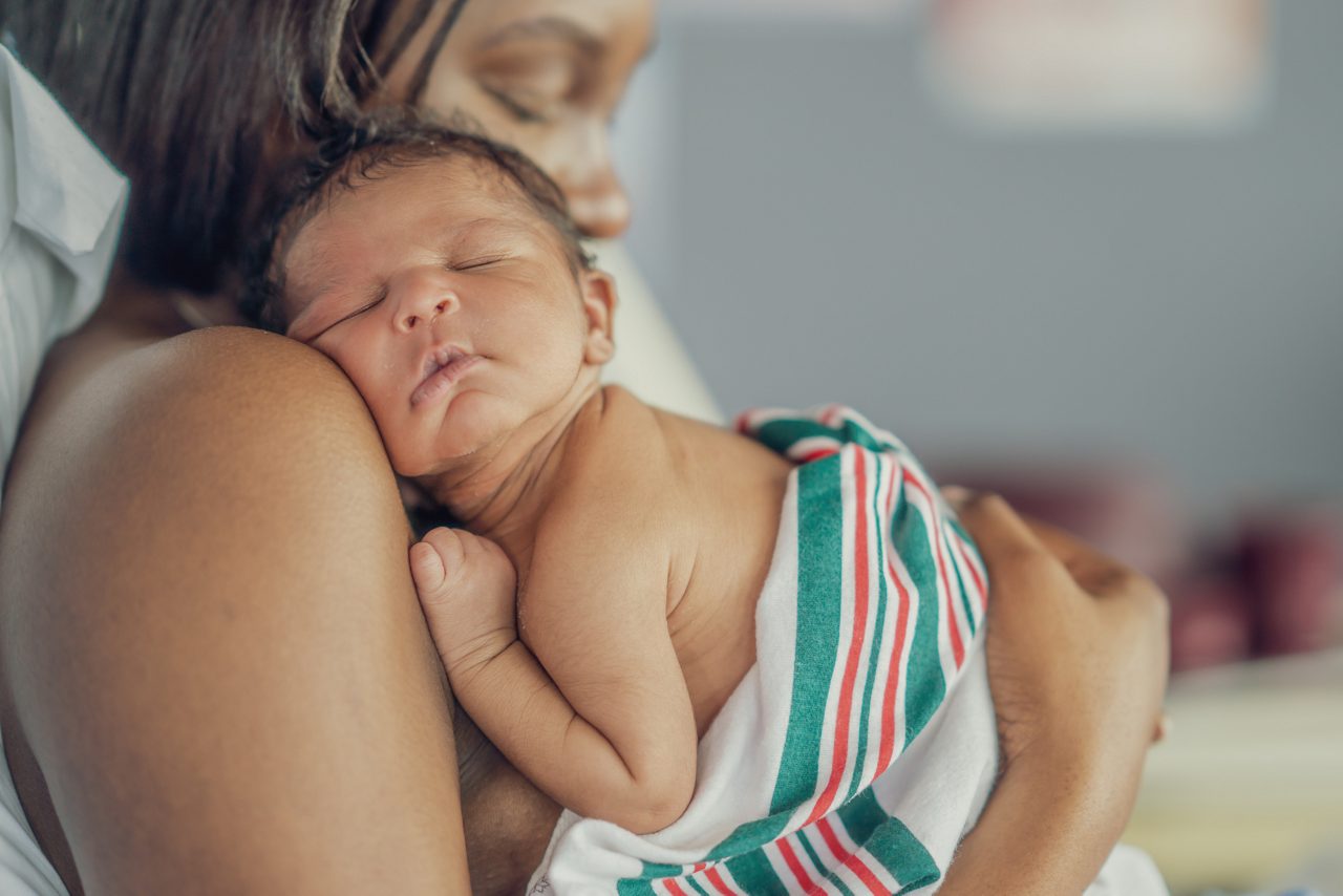A new Mother, of African decent, holds her baby on her shoulder as she sits up in her hospital bed and they spend time bonding skin-to-skin.  The Mother has her eyes closed as she rests from exhaustion after childbirth.
