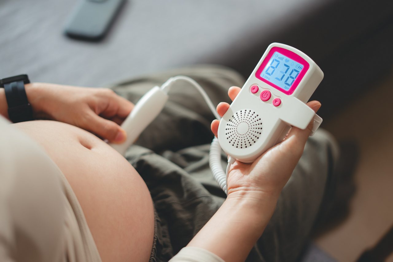 Close-up shot of pregnant woman sitting on sofa in living room at home, using pocket fetal Doppler for listening baby's heartbeat.