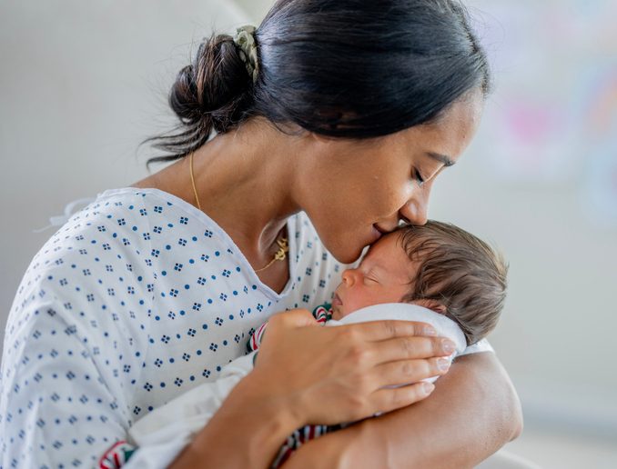 A new Mother sits in  rocking chair shortly after delivery, as she holds her baby tenderly.  She is wearing a hospital gown and the baby is swaddled tightly.