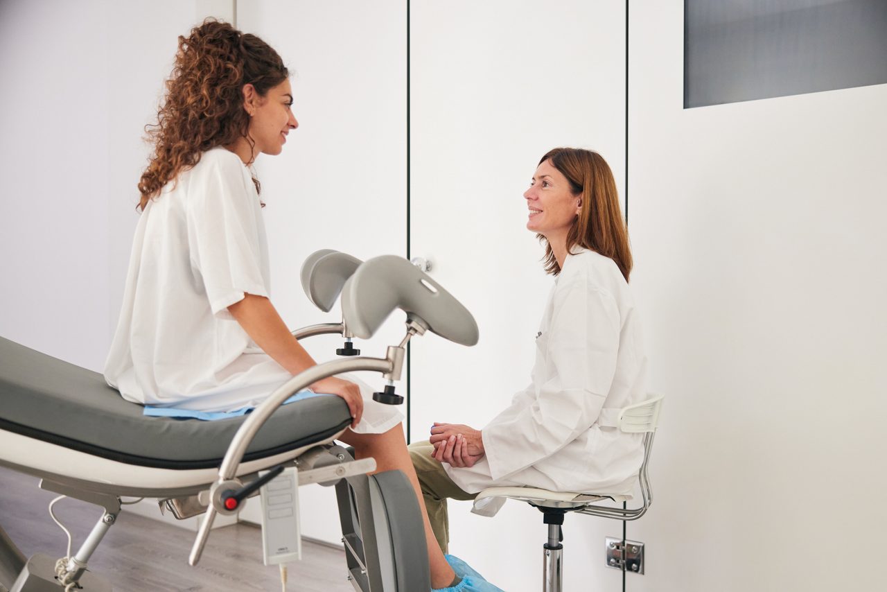 The young woman, dressed in a hospital gown, sits on the stretcher as the mature doctor in a white lab coat sits on a stool, and they engage in discussion, looking at each other.