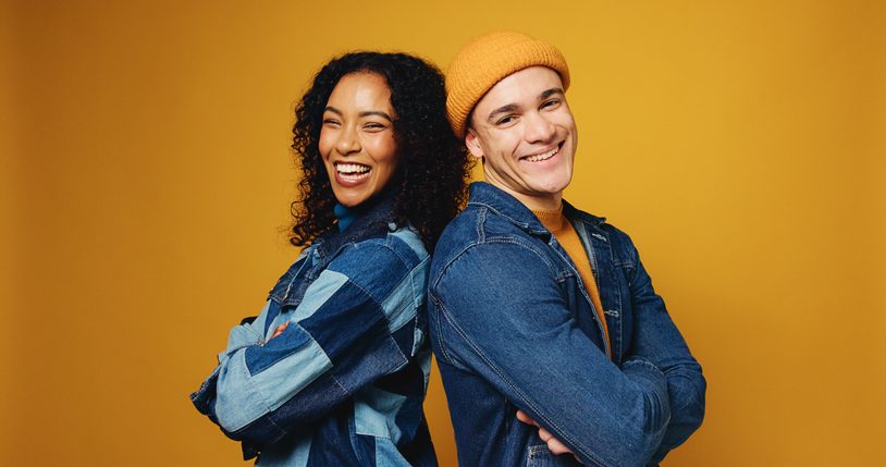Portrait, arms crossed and happy couple in studio for fashion, love and support on yellow background. Man, woman and stylish clothes with confidence, laugh and care for interracial relationship