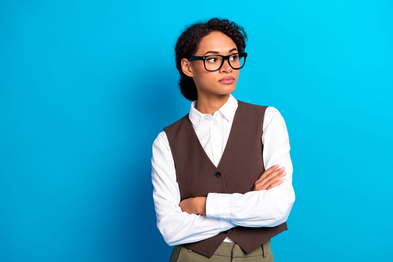 A stylish young female model in white shirt brown vest and green pants stands confidently with arms crossed against a vivid blue background showcasing professional style and modern work vibe.