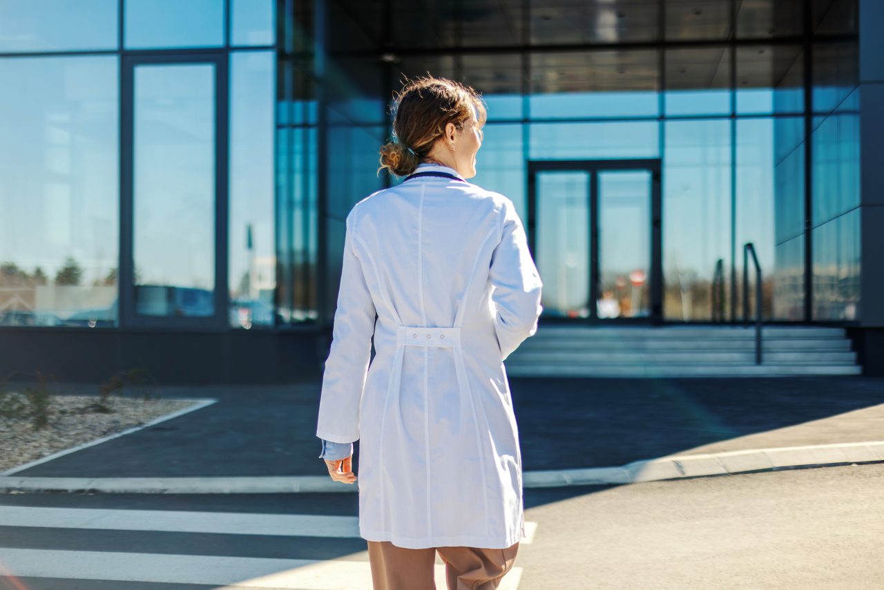 Female doctor in a white lab coat with a stethoscope walking away from the camera, approaching a contemporary glass-fronted healthcare facility on a bright, professional day