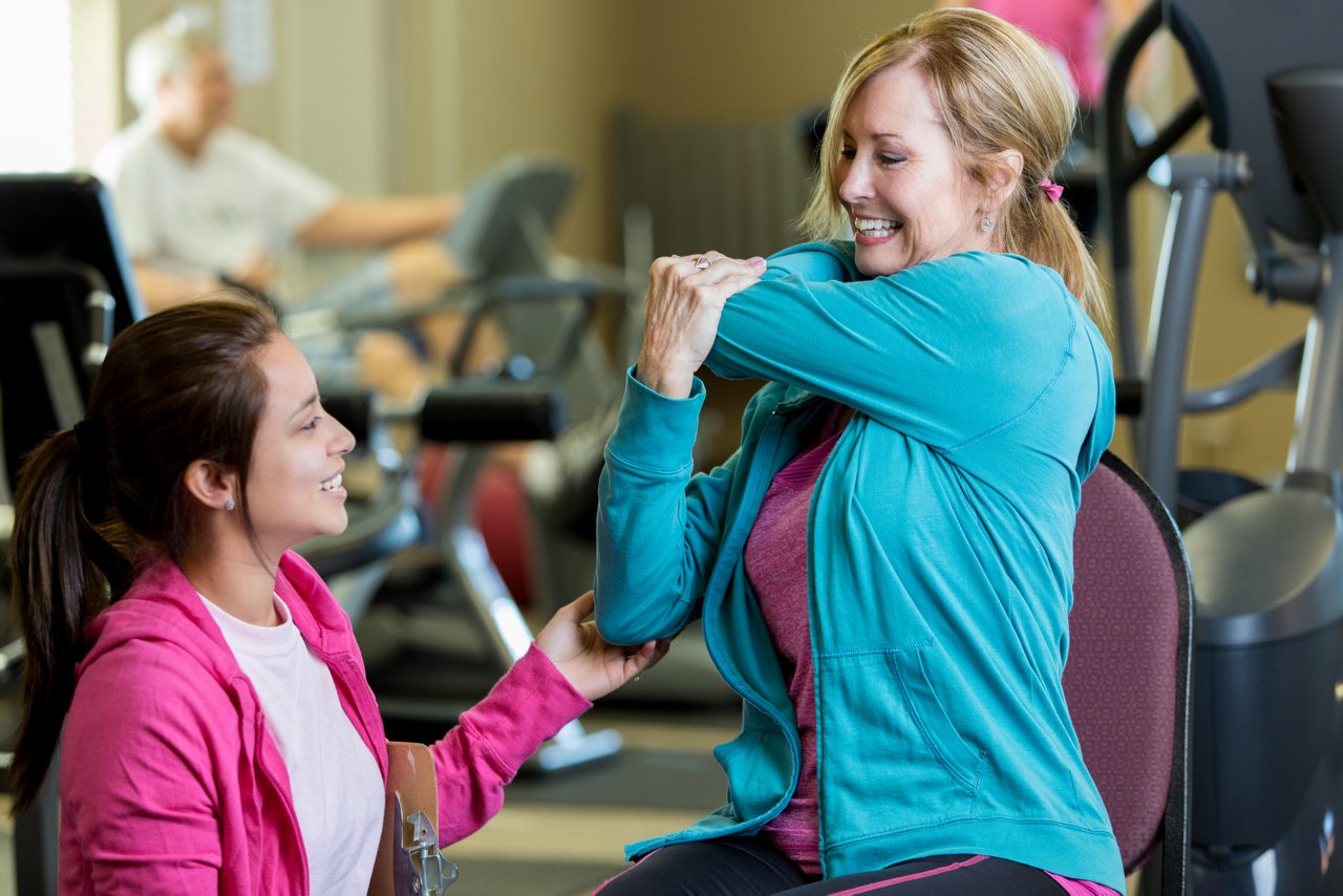 Physical therapist helps senior female client with stretching exercise during therapy approintment. Clients are exercising in the background.