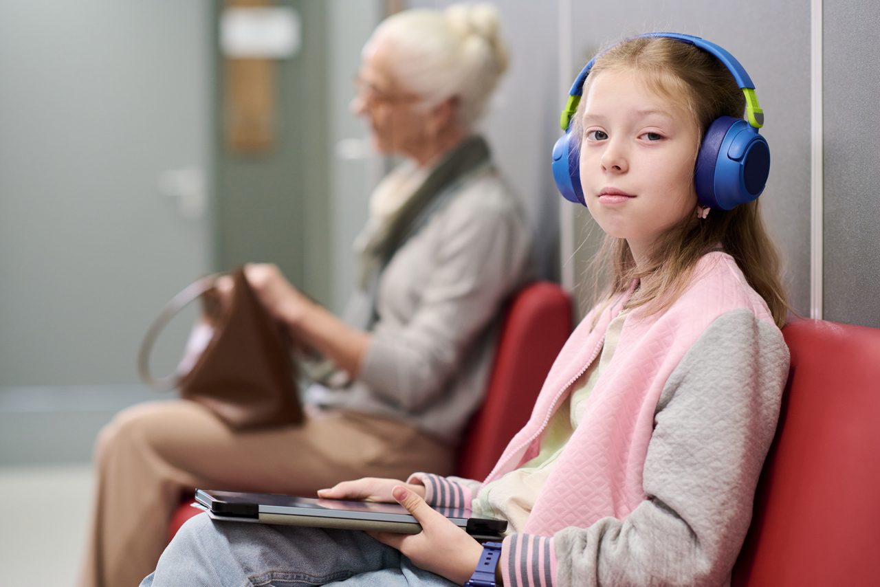 A young girl with headphones on holding a tablet in a waiting room is shown.