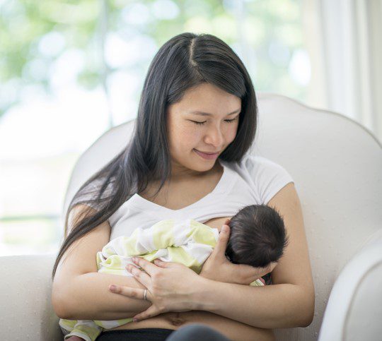 A new Mother sits in rocking chair shortly after delivery, as she holds her baby tenderly. She is wearing a hospital gown and the baby is swaddled tightly.