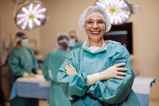 Experienced female surgeon smiles confidently in an operating room while a surgical team works behind her.