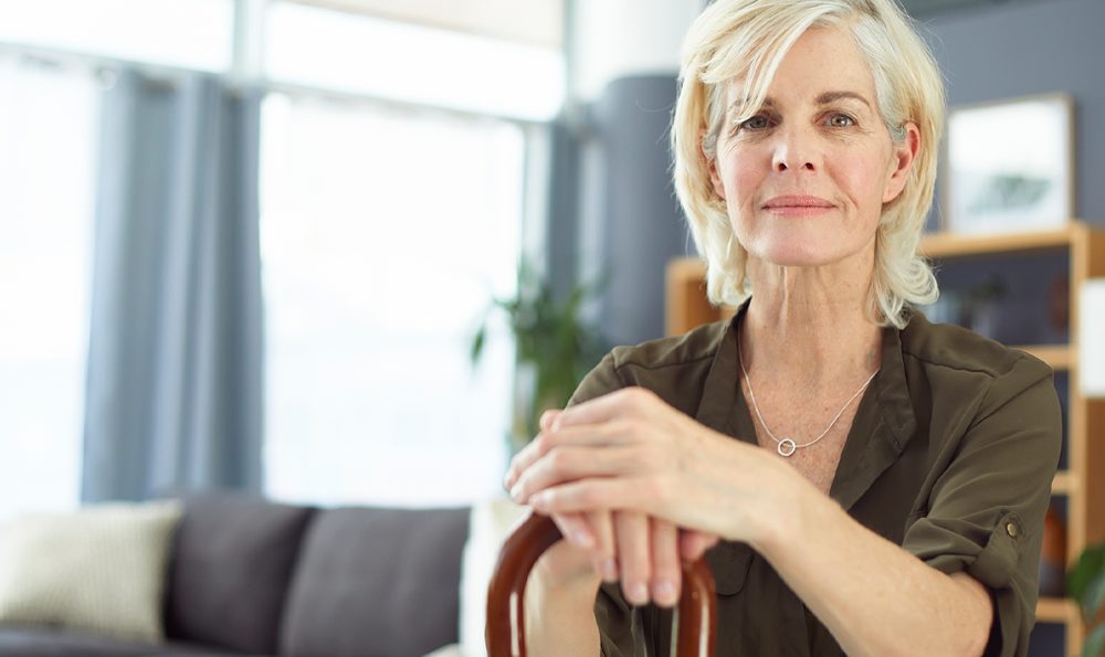 A mature woman sits in her living room, holding a walking cane.