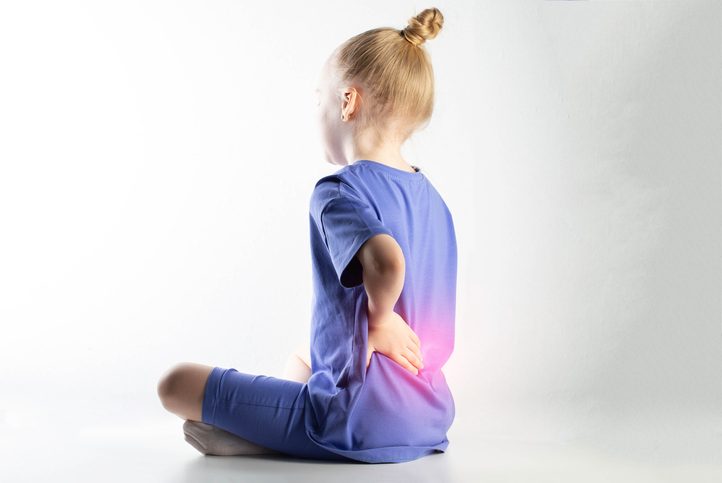 A girl in a purple suit sits and holds her lower back on a white background. Concept of pain and inflammation in the spine in children. Sprains and strains of the back ligaments.