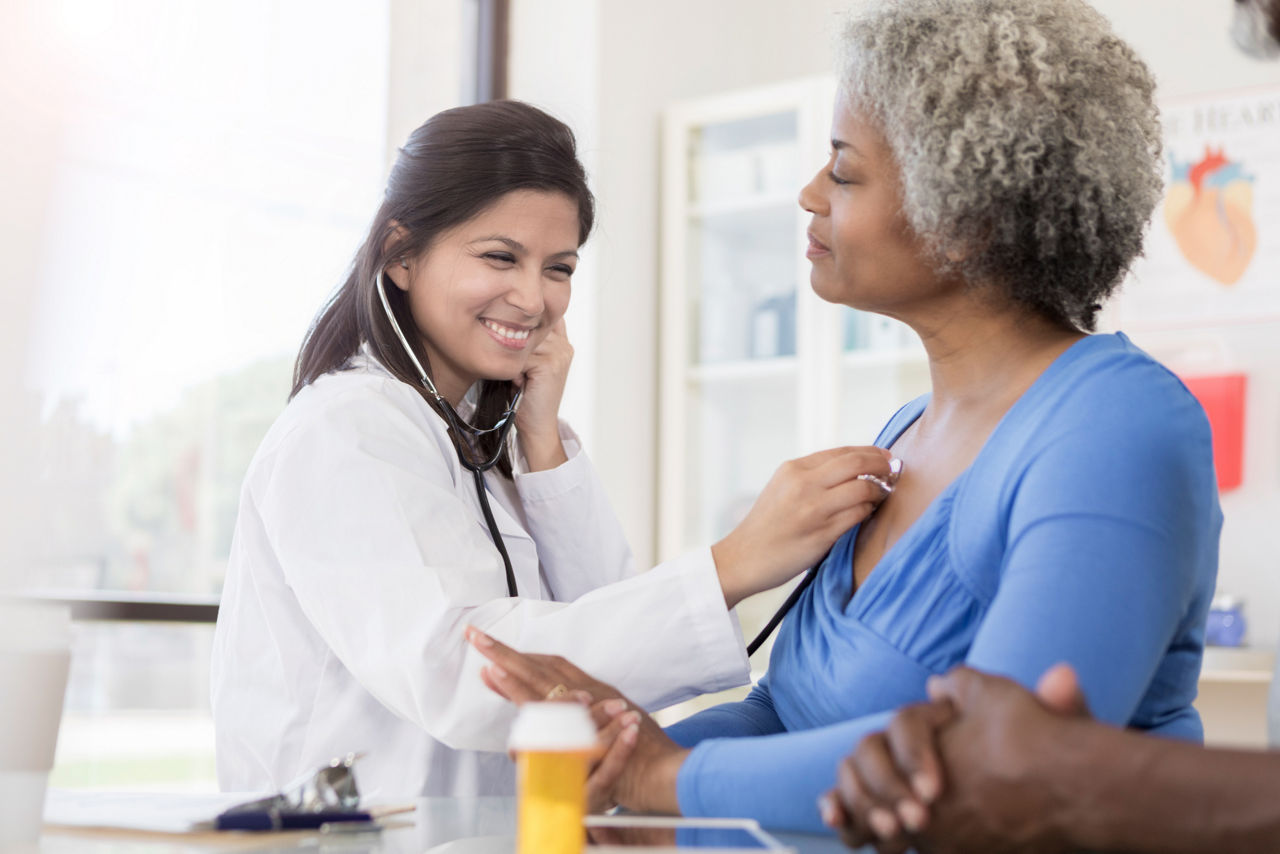 A smiling female doctor sits at a table next to a senior female patient.  She uses her stethoscope to listen to her patient's lungs.  There is a clipboard and prescription bottle on the table.  The hands of another person can be seen resting on the table in the foreground.