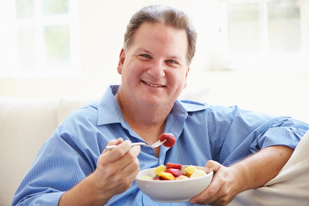 Overweight Man Sitting On Sofa Eating Bowl Of Fresh Fruit At Home Smiling To Camera