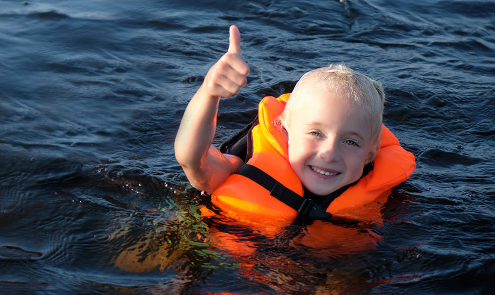 A young child in a life jacket with their thumb up in a body of water.