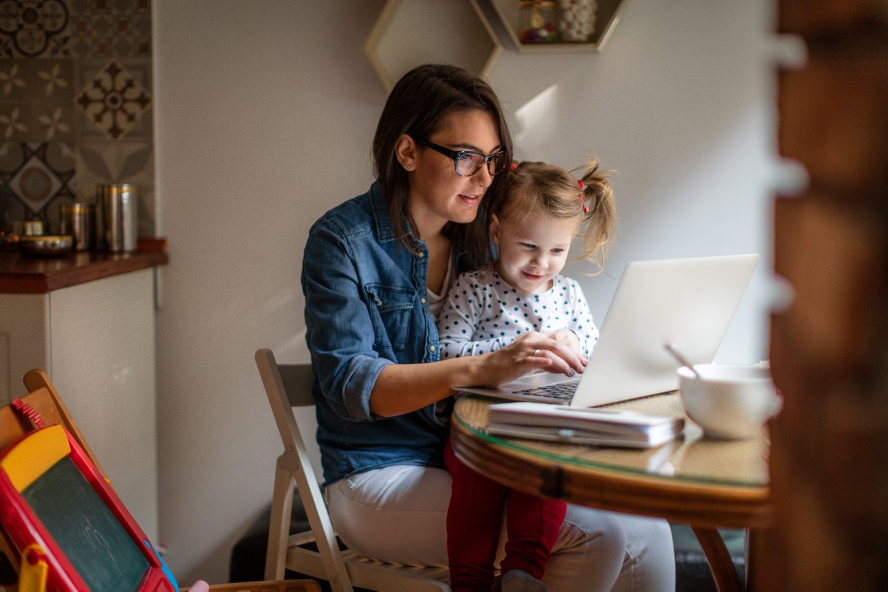 Photo of a little girl trying to help mom who is working on a computer at their dining table